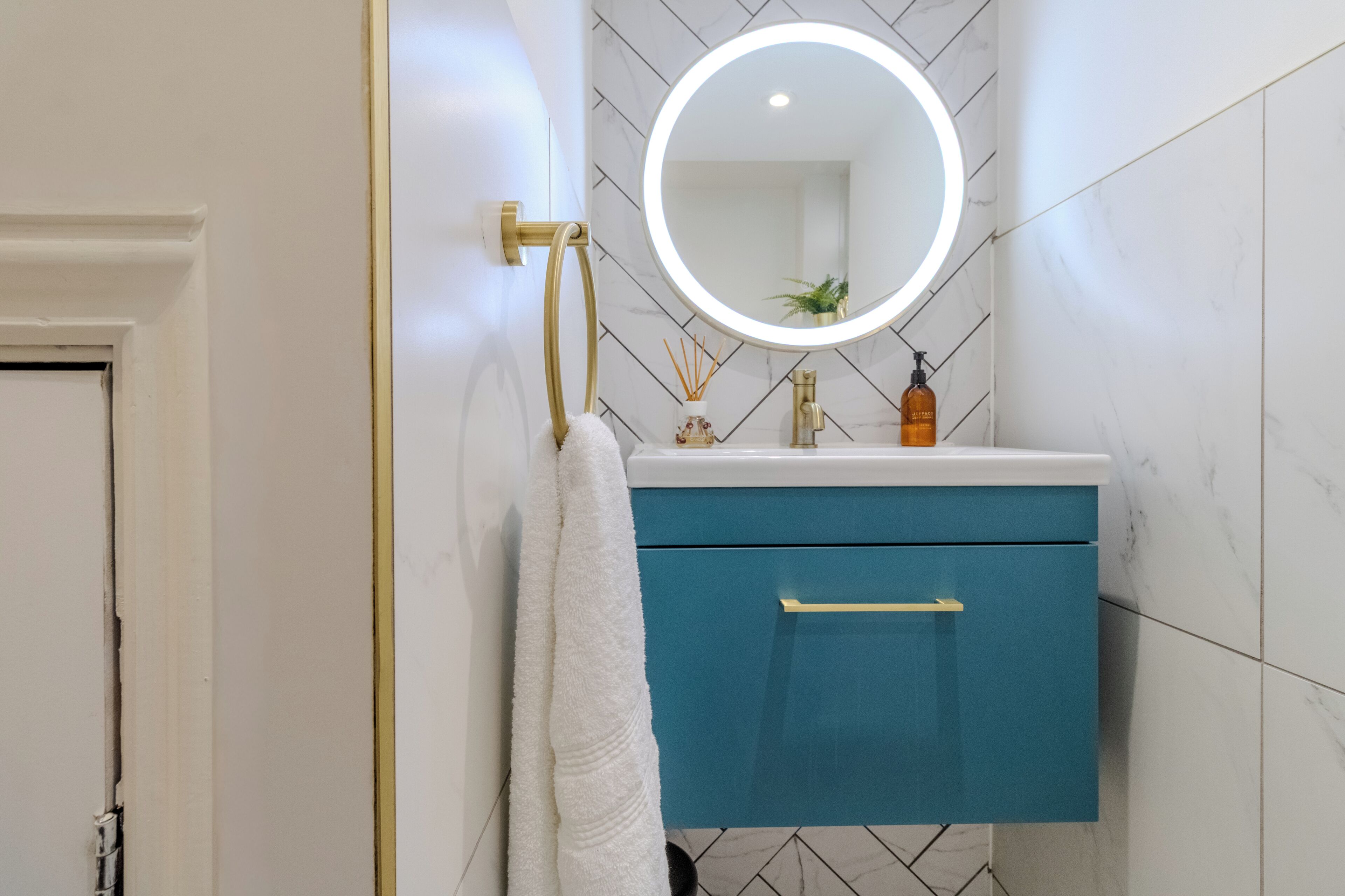A bathroom with a white door, a blue cabinet, a gold towel rack, a round mirror with a light, and a bottle of soap.