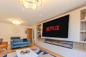 A modern living room with a large flat-screen TV mounted on the wall, a fireplace, a blue sofa, and a coffee table. - Family Friendly 4 Bedroom House in Rayleigh (Essex)