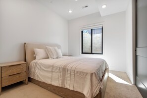 Ein Schlafzimmer mit Bett, Nachttisch und Fenster mit Blick auf eine weiße Wand und eine Mauer.