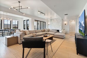 Ein modernes Wohnzimmer mit einem Ecksofa, einem Couchtisch und einem Fernseher. Es gibt einen Essbereich mit Tisch und Stühlen. Der Raum verfügt über große Fenster und einen Blick auf die Berge.