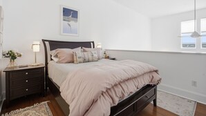 A bedroom with a bed, nightstands, a wall-mounted lamp, a framed picture, and a window. - Brooklyn Arts Condo | Walk to Live Oak & Dining (Wilmington)
