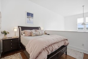 Ein Schlafzimmer mit einem Bett, Nachttischen, einer Wandlampe, einem gerahmten Bild und einem Fenster.
