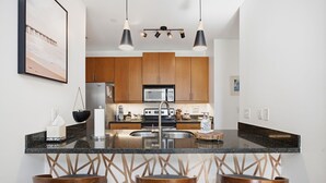 A modern kitchen with a central island, bar stools, wooden cabinets, stainless steel appliances, and pendant lighting. - Brooklyn Arts Condo | Walk to Live Oak & Dining (Wilmington)