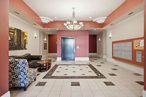 En hotellobby med en kristallkrona, en soffa, en mönstrad pall, ett soffbord, en tavla, en dörr och en anslagstavla med förvaringsskåp.