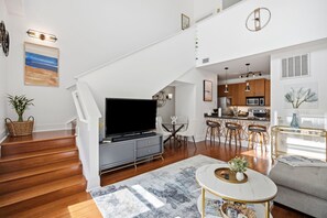 Ein modernes Wohnzimmer mit Flachbildfernseher, einem Essbereich mit Glastisch und einer Küche mit Holzschränken.