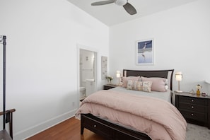 A bedroom with a bed, bedside tables, a ceiling fan, and a door leading to a bathroom. - Brooklyn Arts Condo | Walk to Live Oak & Dining (Wilmington)