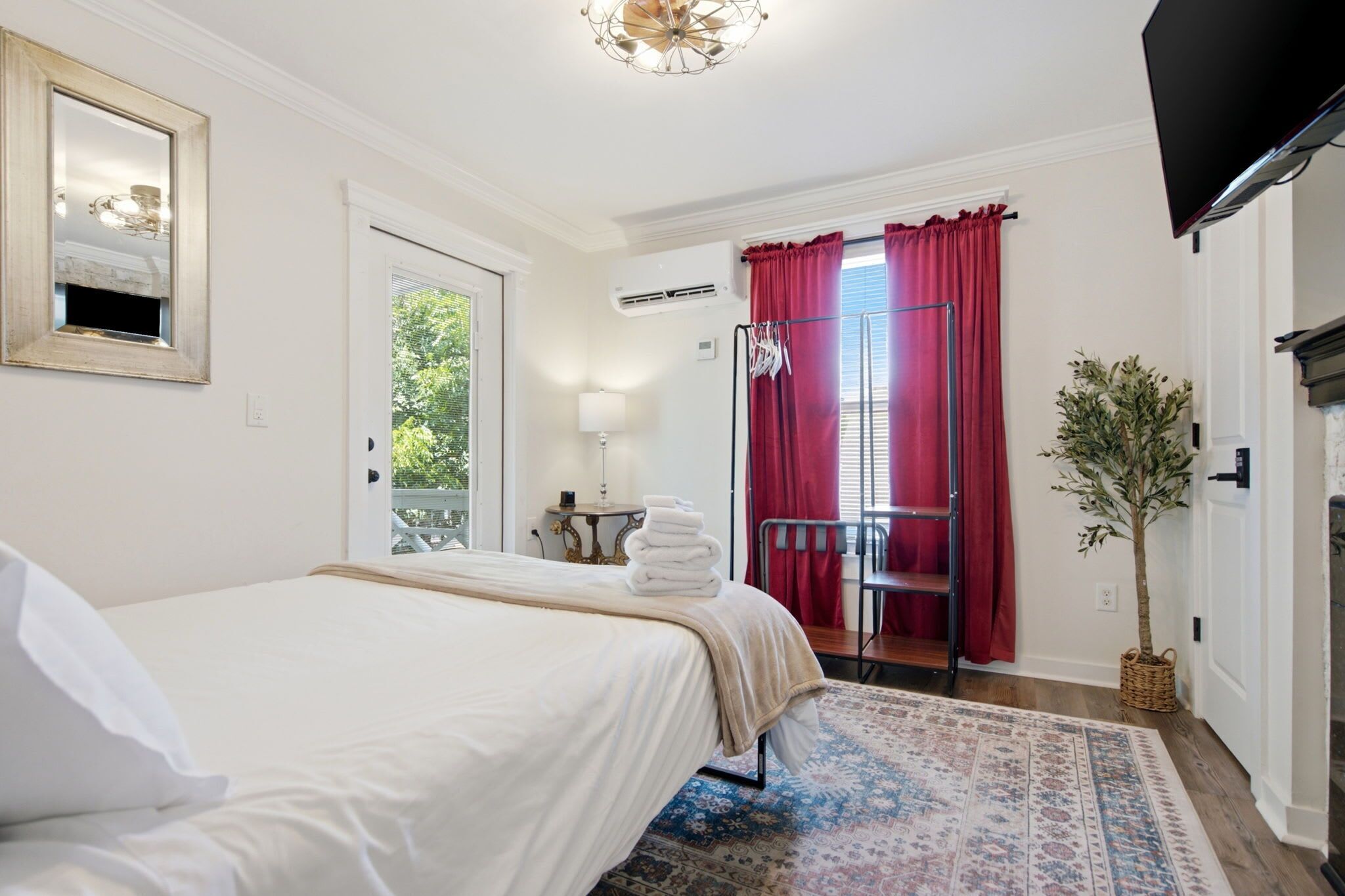 A hotel room with a bed, a small table with a lamp, a window with curtains, a radiator, and a plant.