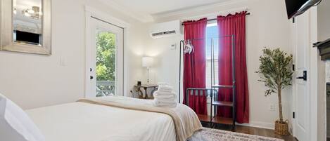 A hotel room with a bed, a small table with a lamp, a window with curtains, a radiator, and a plant.