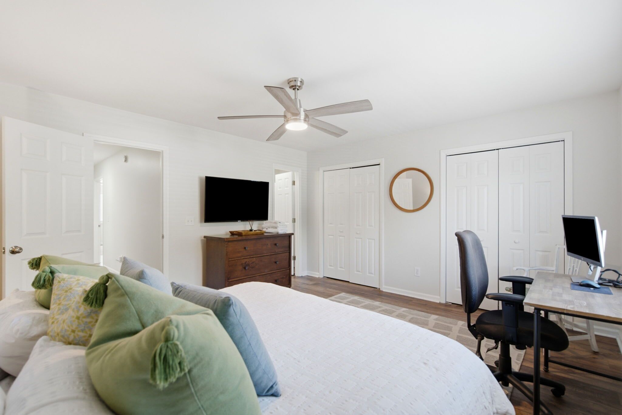 Une chambre à coucher comprenant un lit, une commode, un téléviseur, un bureau avec un ordinateur, une chaise et un ventilateur de plafond.