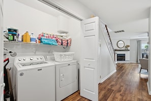 A laundry area with a washing machine, dryer, and a shelf with folded towels and laundry supplies. - Minutes to Wrightsville Beach, Downtown & UNCW (Wilmington)