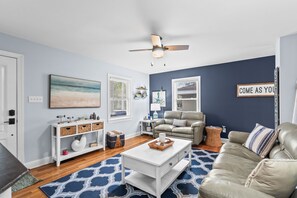 Un salon avec une table basse blanche, un canapé beige, une table d’appoint blanche et un ventilateur de plafond.