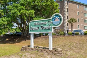 A welcome sign for Sunset Beach with a view of a building and parked cars. - Cozy Beach Cottage Near Ocean Isle Beach (Ocean Isle Beach)