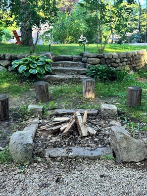 Ein Steinkamin, umgeben von Steinen und Holzstämmen, in einer grünen Fläche mit einer Steinmauer und Pflanzen im Hintergrund.