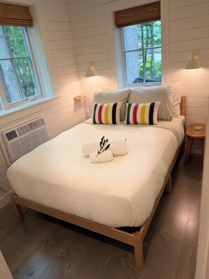 Een netjes opgemaakt bed met een witte dekbedovertrek, kussens en een handdoek met een bloemenmotief. Er staat een nachtkastje met een lamp, een raam met uitzicht op bomen, en een wandradiator.