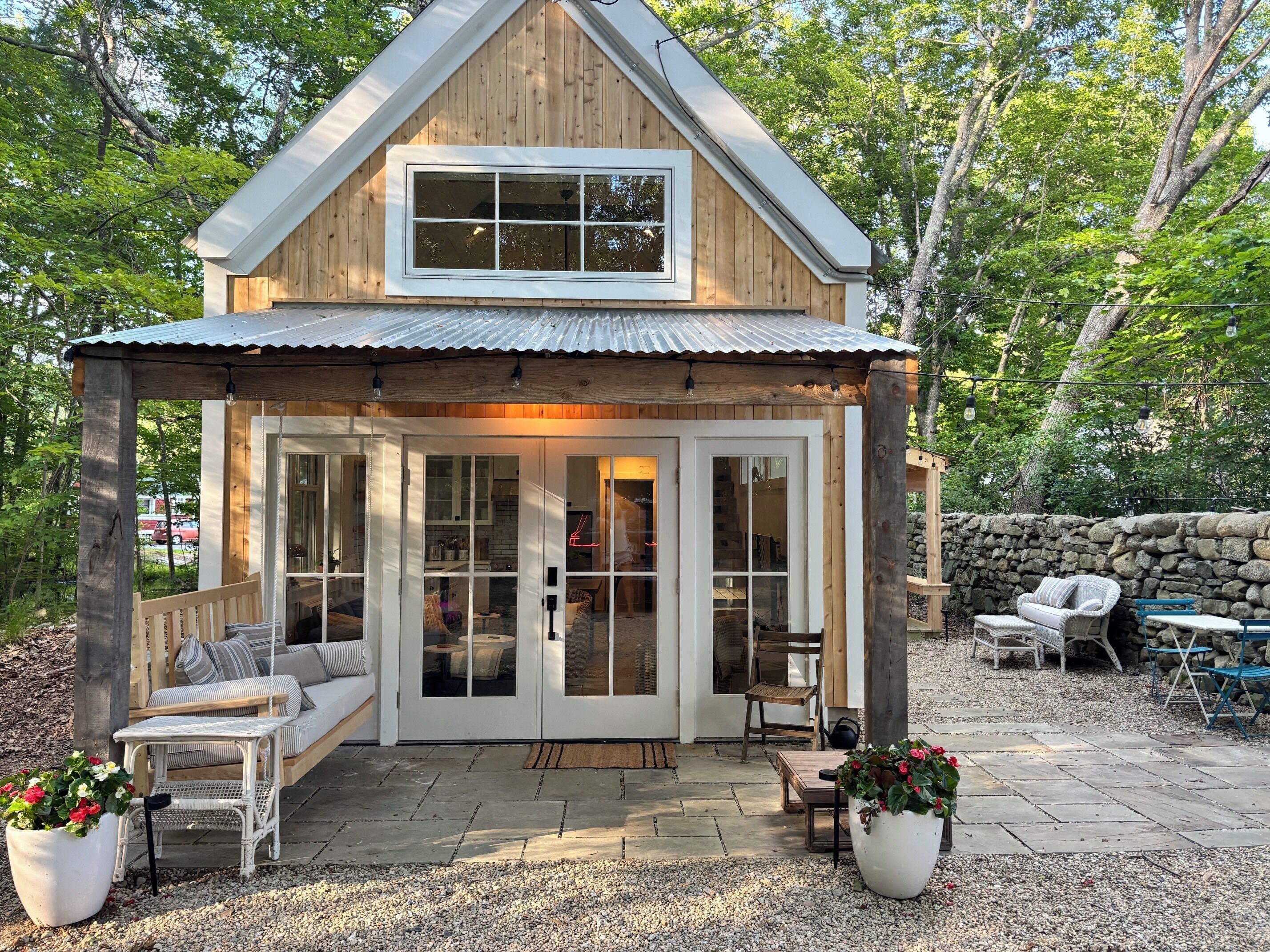 A wooden cabin with a porch, large windows, and a stone wall in the background.