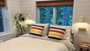 Ein Schlafzimmer mit einem großen Bett, zwei Nachttischlampen, einem Fenster mit Blick auf Bäume und einer Zimmerpflanze.