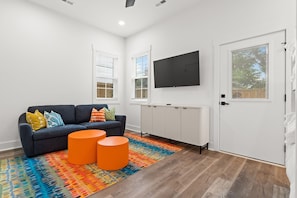 A modern living room with a dark grey sofa, colorful cushions, two orange side tables, a large flat-screen TV, and a radiator. - Rooftop Spa + Bar | Walk to Beach | Sleeps 15! (Carolina Beach)