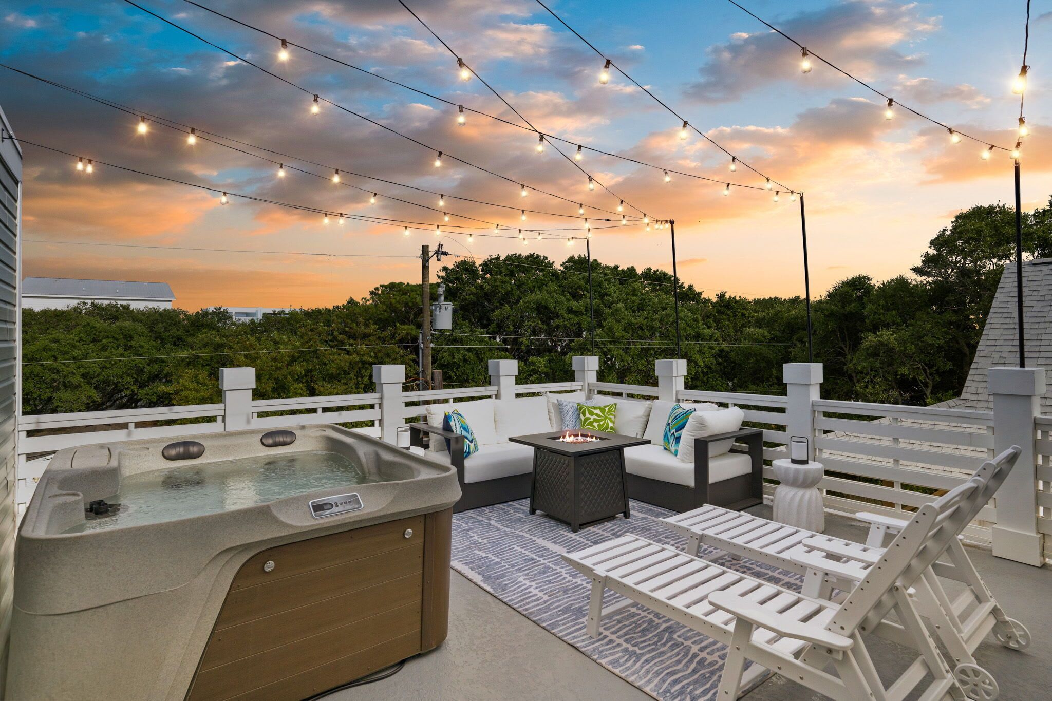 An outdoor patio with a hot tub, seating area, and string lights against a sunset backdrop.