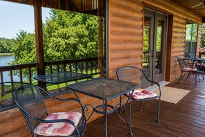 Eine Holzterrasse mit Gartenmöbeln, eine Glastür, die in einen überdachten Bereich führt, und Blick auf einen See.