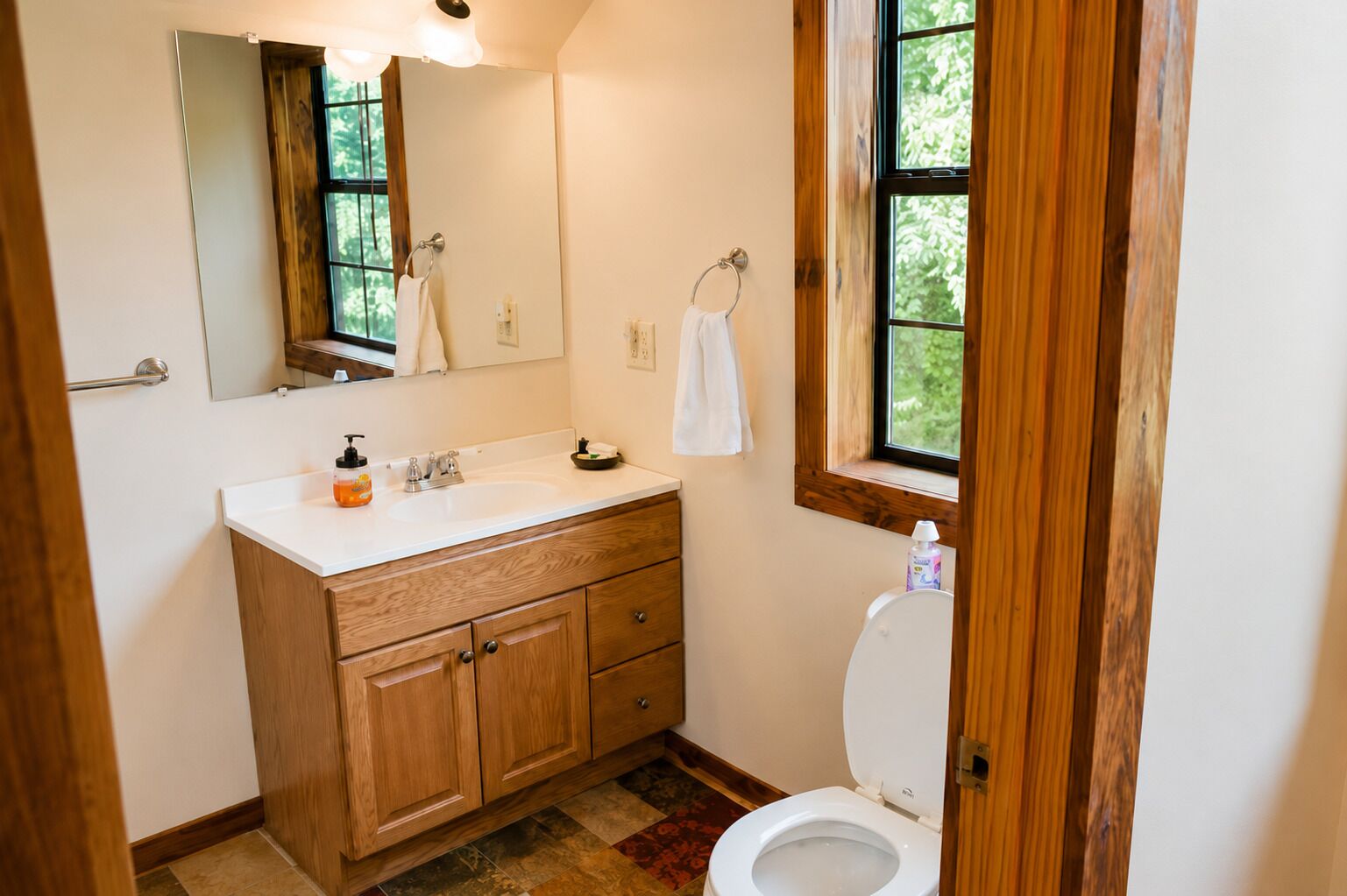 Een badkamer met een houten wastafelmeubel, een witte wastafel, een spiegel, een toilet en een raam met uitzicht op bomen.