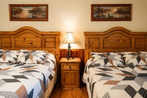 Ein Zimmer mit zwei Betten, jedes mit einer gemusterten Steppdecke, einem Nachttisch mit Lampe sowie zwei gerahmten Landschaftsbildern an der Wand.
