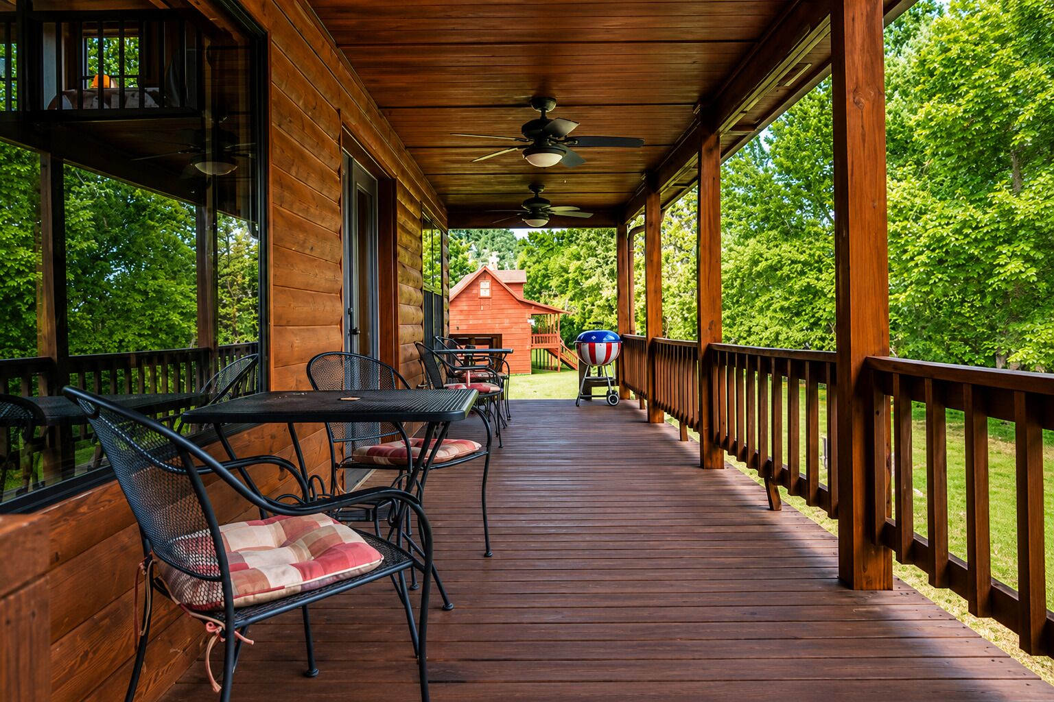 A covered wooden deck with outdoor seating, a barbecue grill, and a view of a red building in the distance.