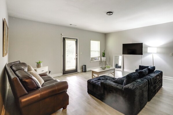 Ein Wohnzimmer mit einem braunen Ledersofa, einem schwarzen gewebten Hocker, einem weißen Beistelltisch mit Pflanze und einem Fernseher auf einem Schrank.