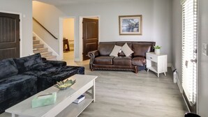 A living room with a dark brown leather sofa, a white coffee table, and a white side table with a plant. A staircase is visible in the background leading to another room. - Shop, Dine & Hike: Lawrenceville Home By Parks (Lawrenceville)