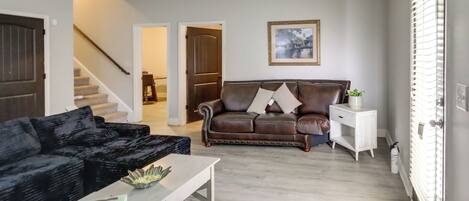 A living room with a dark brown leather sofa, a white coffee table, and a white side table with a plant. A staircase is visible in the background leading to another room.