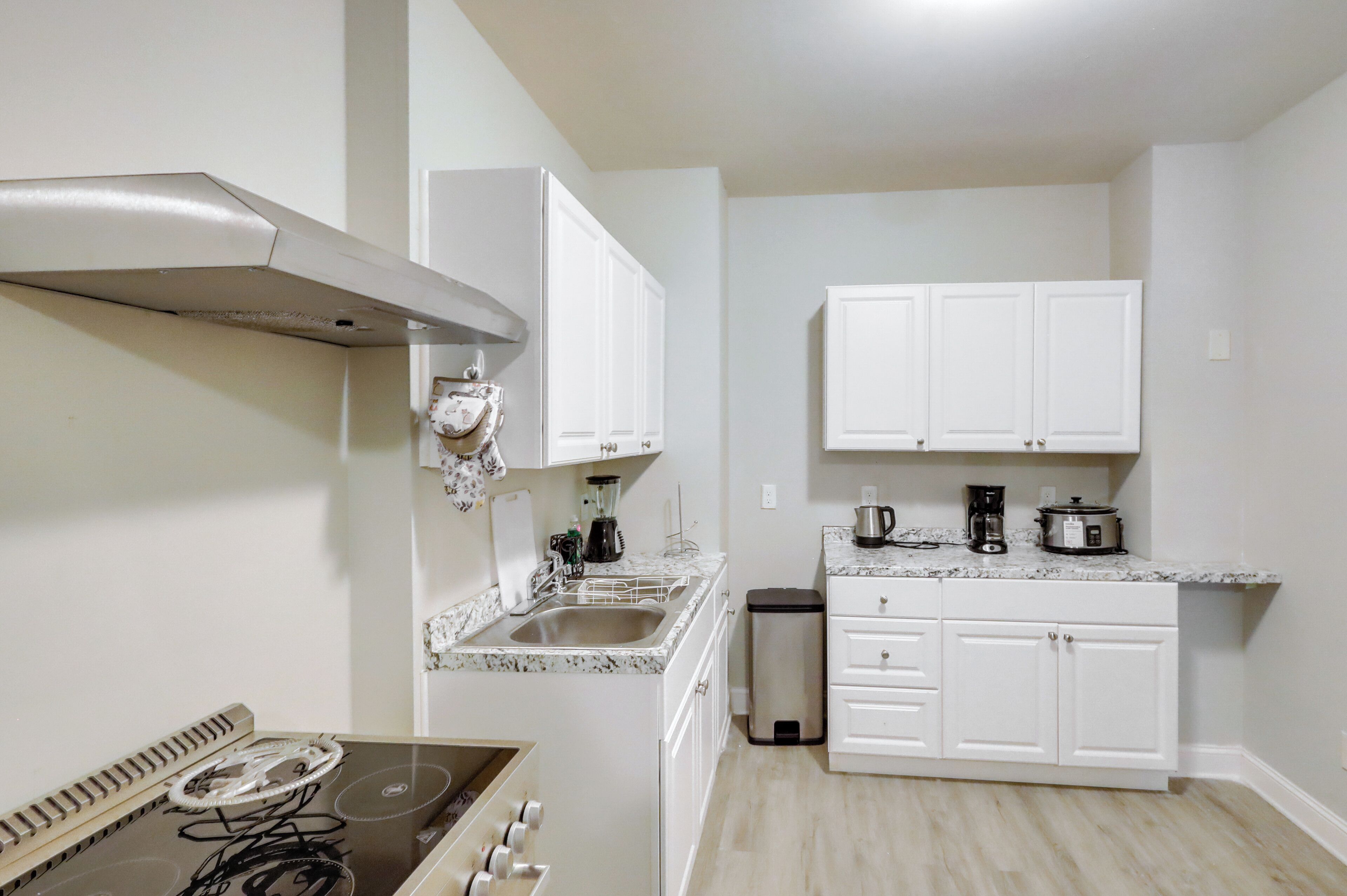 A kitchen with white cabinets, a stainless steel range hood, a sink, and a countertop with a coffee maker and a slow cooker.