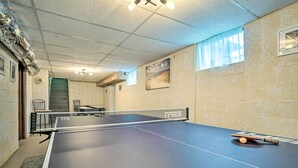 Una mesa de ping pong en una habitación con techo texturizado, una ventana con cortina translúcida y un cuadro en la pared.