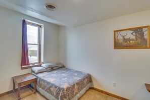 Un dormitorio con cama, ventana con cortinas, una mesita, un cuadro en la pared y piso alfombrado.