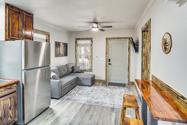 A compact kitchen with a stainless steel refrigerator, a gray sectional sofa, and a wooden dining table.