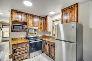 A compact kitchen with wooden cabinets, stainless steel appliances including a microwave, stove, and refrigerator, and a window with blinds.