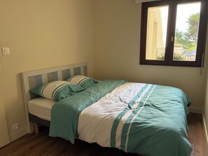 Ein Bett mit gestreifter Bettdecke und Kissen, ein Fenster mit Blick ins Grüne und ein Lichtschalter an der Wand.