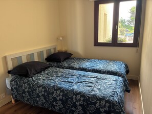 A bedroom with two single beds, a nightstand, a lamp, and a window with a view of the outside. - Fully renovated apartment with direct access to the beach (La Turballe)