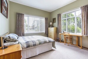 Ein Schlafzimmer mit einem Bett, einem Schreibtisch, einem Sessel, einer Kommode und einem Fenster mit Vorhängen.