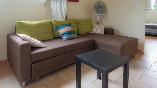 Una sala de estar con un sofá marrón, almohadas verdes y multicolores, una pequeña mesa negra y un ventilador de pie.