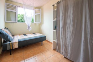 A single bed with a patterned bedspread, a window with a sheer curtain, and a white shower curtain. - Le Lamentin - Patio - TV (Le Lamentin)