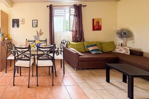 A living room with a sofa, a dining table with chairs, a fan, and a window with curtains. - Le Lamentin - Patio - TV (Le Lamentin)