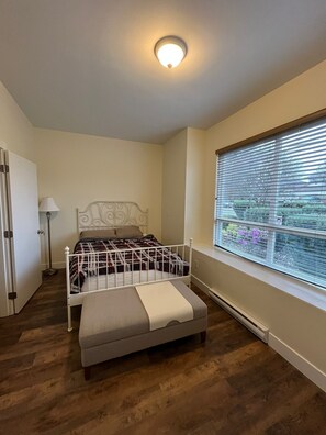 A bedroom with a bed, a bedside table, a lamp, a window with blinds, and a radiator. - Modern & Cozy Stay | Self Check-In | Free Parking (Surrey)