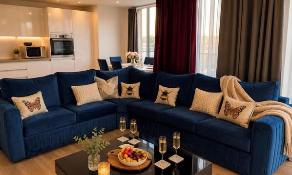A modern living room with a blue sectional sofa, a coffee table with fruit and glasses, and a kitchen in the background. - Southampton 2-bed Apartment, Sleeps 4 With Balcony (Southampton)