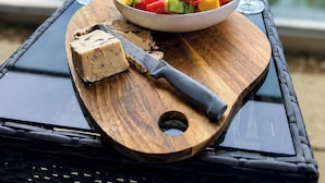 Ein Holzboden mit Obstsalat, zwei Gläsern Weißwein und einem Messer auf einem Tisch mit Blick auf ein Gewässer.