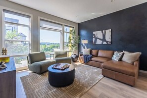 A modern living room with a large window, a brown leather sofa, a grey armchair, a round coffee table with chess pieces, and a dark wall with a painting. - Luxury Boutique Stay w/ Hot Tub | Sleeps 8 | Near Golf Courses (Edmonton)
