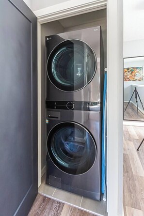 A compact, modern laundry room with a built-in washer and dryer, a small shelf, and a tripod in the background. - Luxury Boutique Stay w/ Hot Tub | Sleeps 8 | Near Golf Courses (Edmonton)