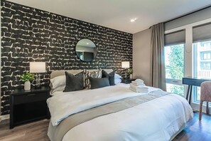 Ein modernes Schlafzimmer mit einer Wand mit Muster, einem runden Spiegel, einem Bett mit weißer Bettwäsche und Blick auf Grün durch das Fenster.