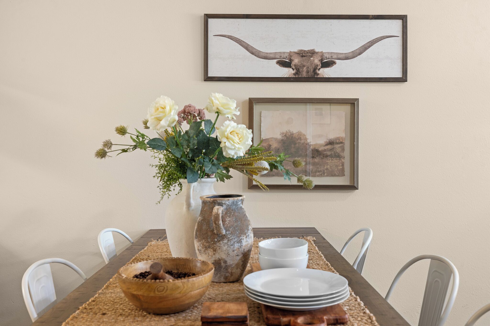 Uma mesa de jantar posta com pratos brancos, uma tigela de madeira, um jarro de cerâmica e um vaso com flores.