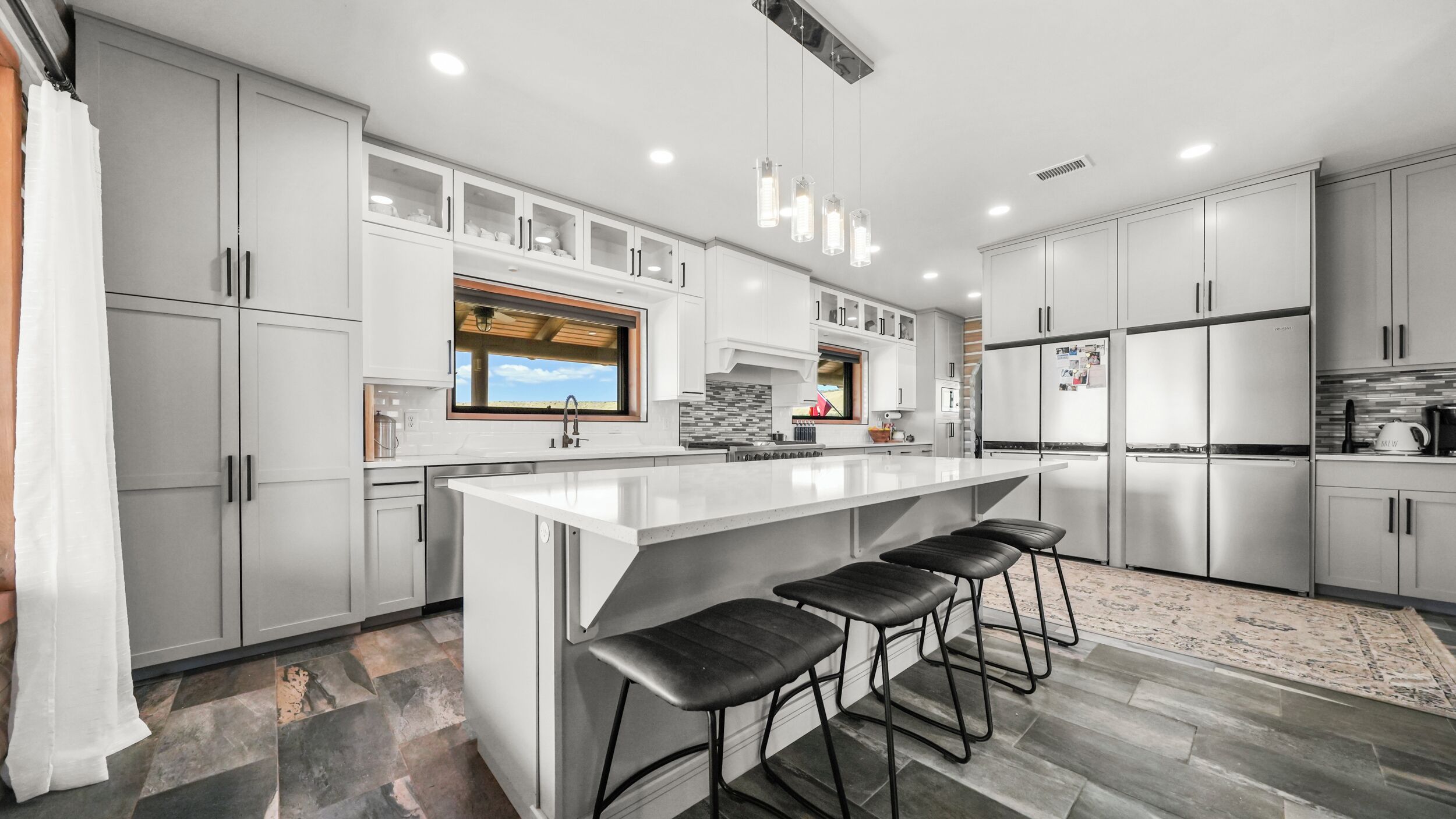 A modern kitchen with white cabinetry, a large island with bar stools, and a view of the outdoors through the window.