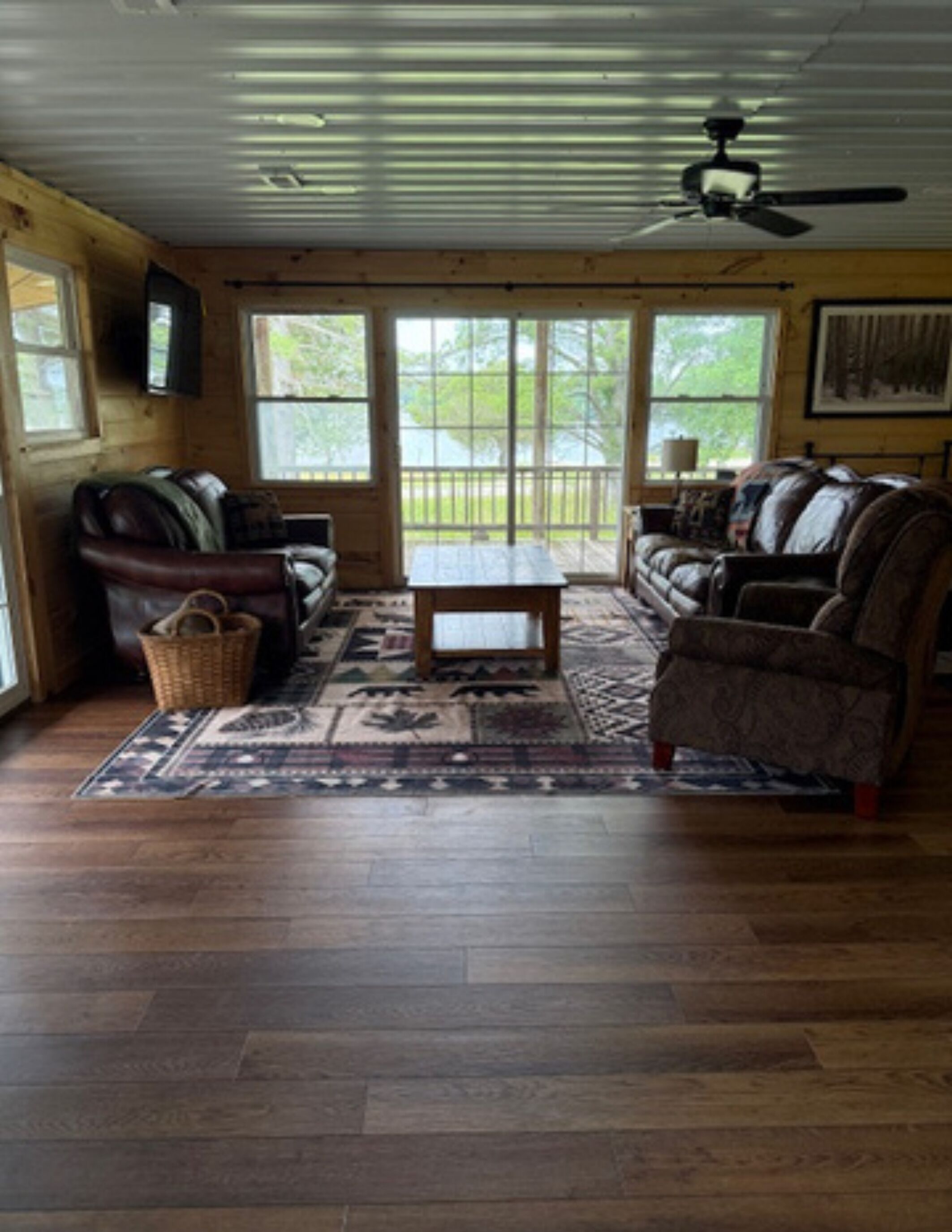 A spacious living room with a wooden floor, a ceiling fan, and a large window with a view of greenery.