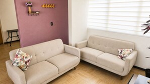 Una sala de estar con dos sofás color beige, una mesita, un gancho en la pared y una ventana con persianas.